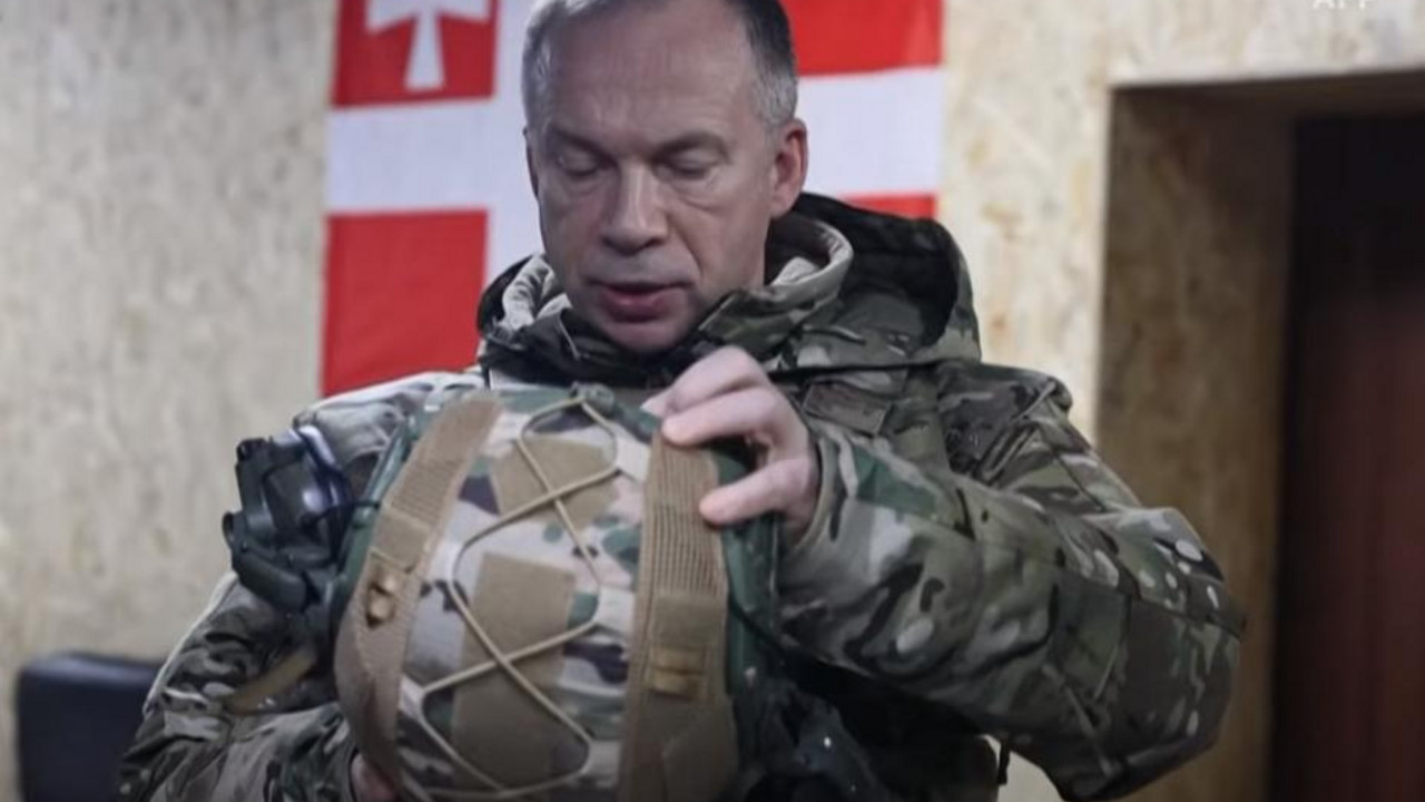 Aleksandar Sirski u uniformi gleda u unutrašnjost šlema dok se iza njega nalazi zastava Gruzije